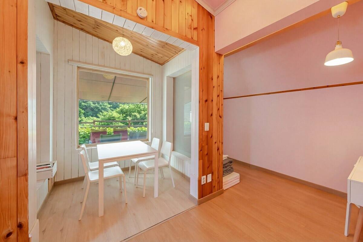 A room with a wooden floor, a white table with chairs, a small white desk, and a window with a view of greenery.