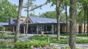 A modern building with a covered outdoor seating area, surrounded by greenery and trees. - Cozy home in a holiday park with a swimming pool (Lage Mierde)