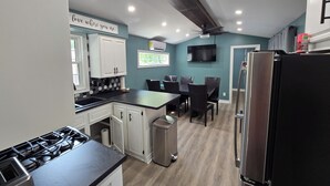 A modern kitchen with white cabinets, a black countertop, a stainless steel refrigerator, and a dining area with chairs and a table. - Freshly remodeled premium trailer in prime location! (Port Colborne)