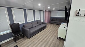 A room with a grey sofa, a black chair, a small desk, and a television on a white wall. - Freshly remodeled premium trailer in prime location! (Port Colborne)