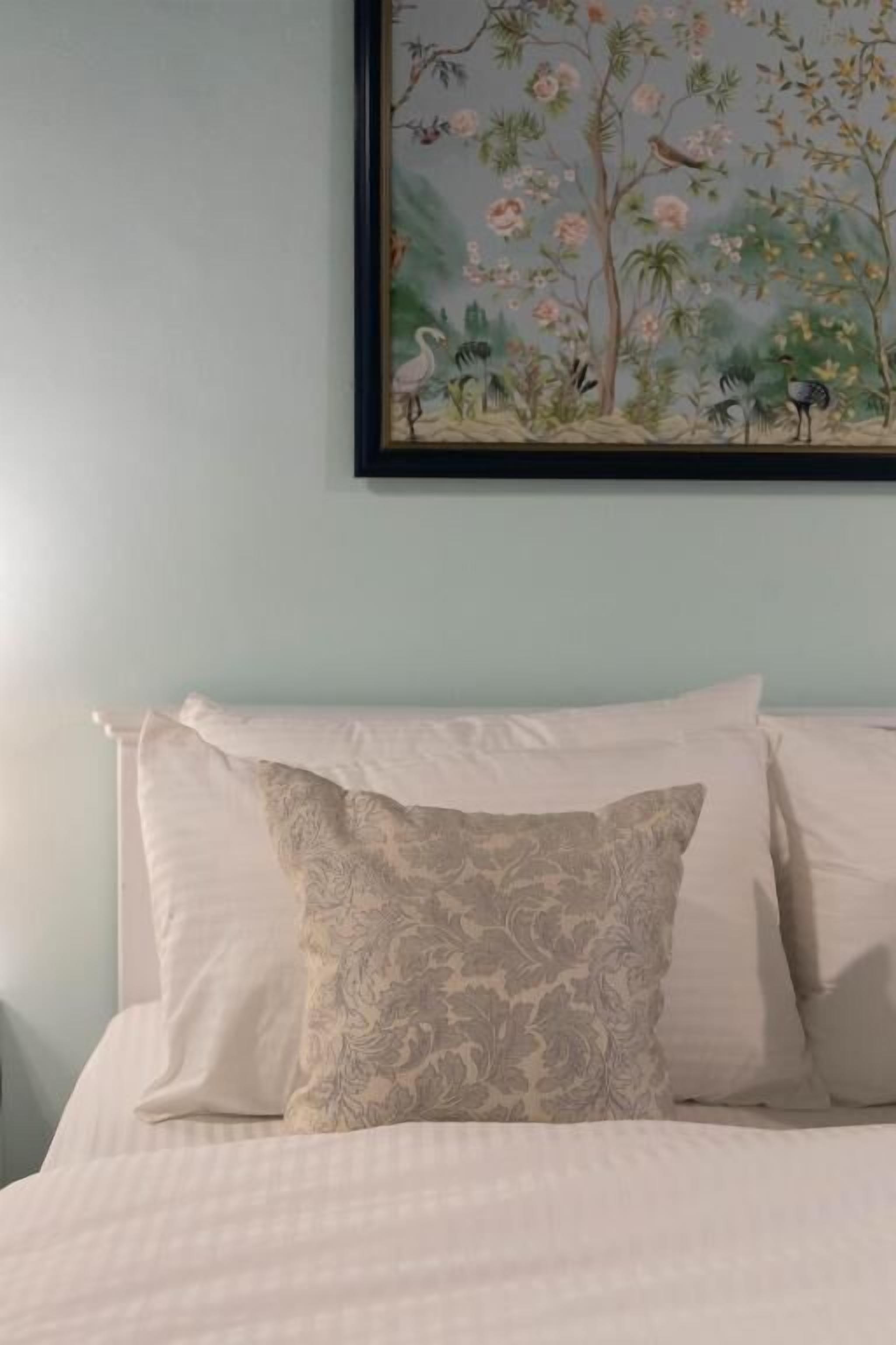 A bed with white bedding and a floral-patterned pillow, a framed botanical painting above the bed, and a light blue wall.