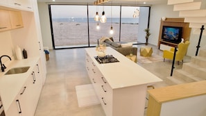 A modern kitchen with a large island, a dining area with yellow chairs, a TV, and a view of the beach. -  Silver Sands Beach Villa (Langstrand)