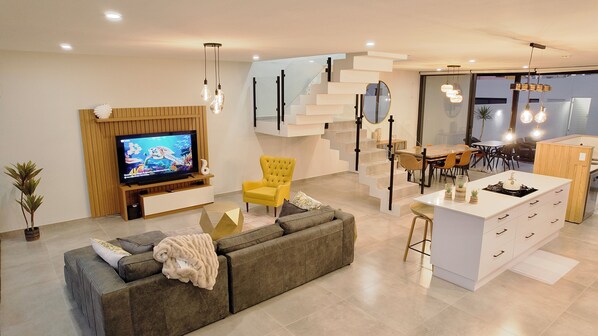 A modern living room with a sofa, TV, yellow armchair, staircase, dining area, and kitchen island.