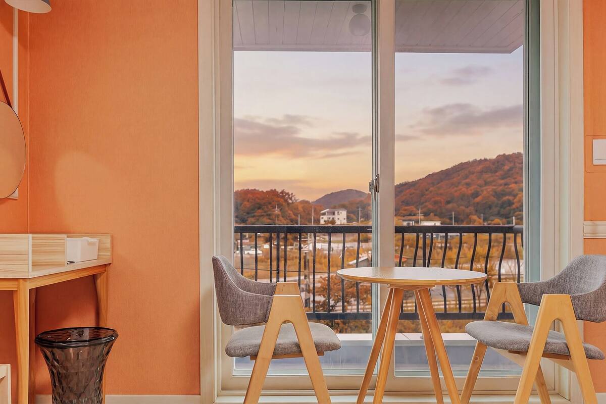 Une pièce avec une table en bois, deux chaises grises, un petit bureau en bois, un vase en verre et une grande fenêtre donnant sur un paysage vallonné.