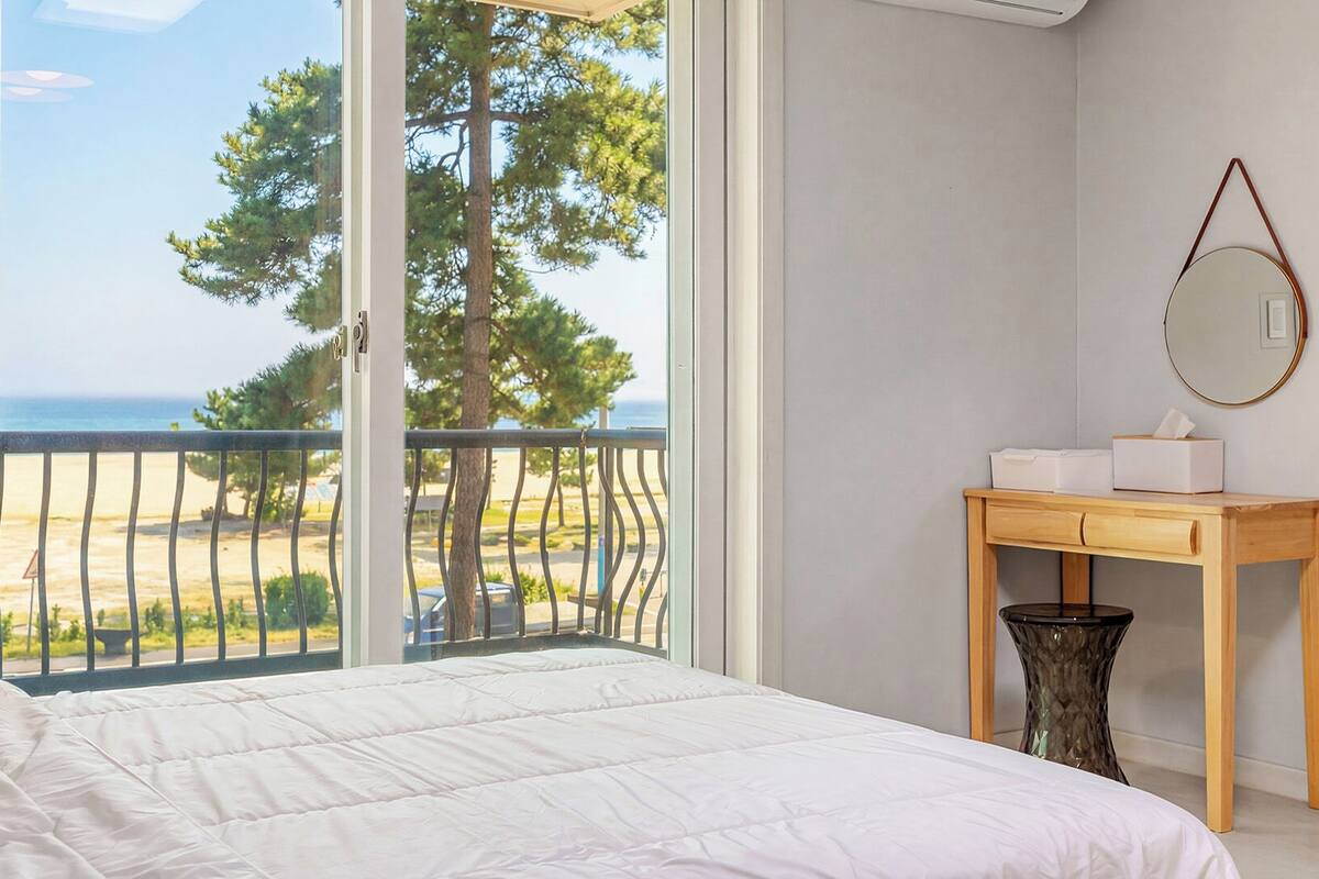A bedroom with a bed, a wooden nightstand, a mirror, and a view of the beach and trees.