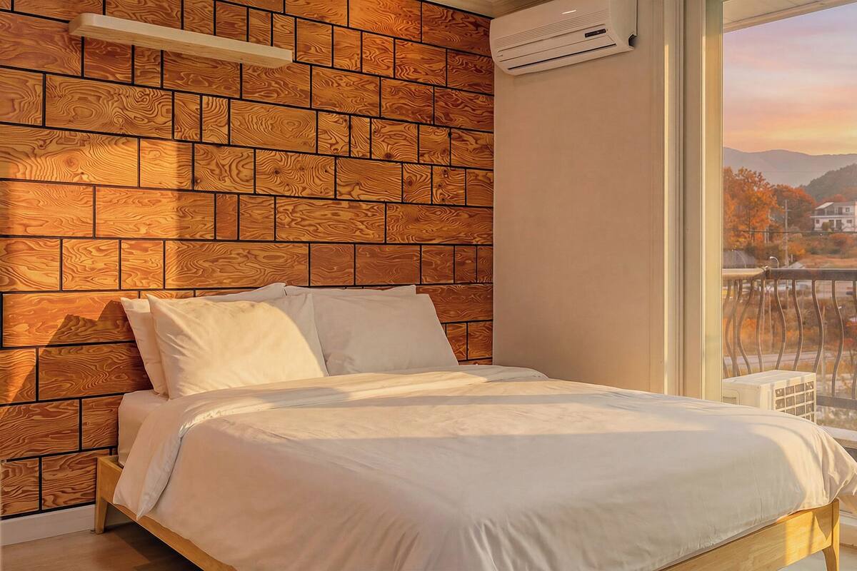 Une chambre avec un mur décoré d’un motif en briques, un lit recouvert de linge de lit blanc et une grande fenêtre donnant sur un paysage urbain.