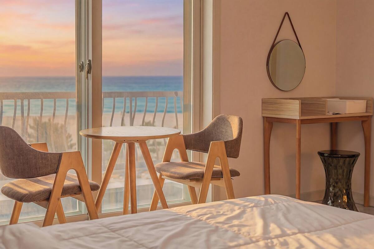 A room with a bed, a round table with two chairs, a small table with a mirror, and a view of the sea.