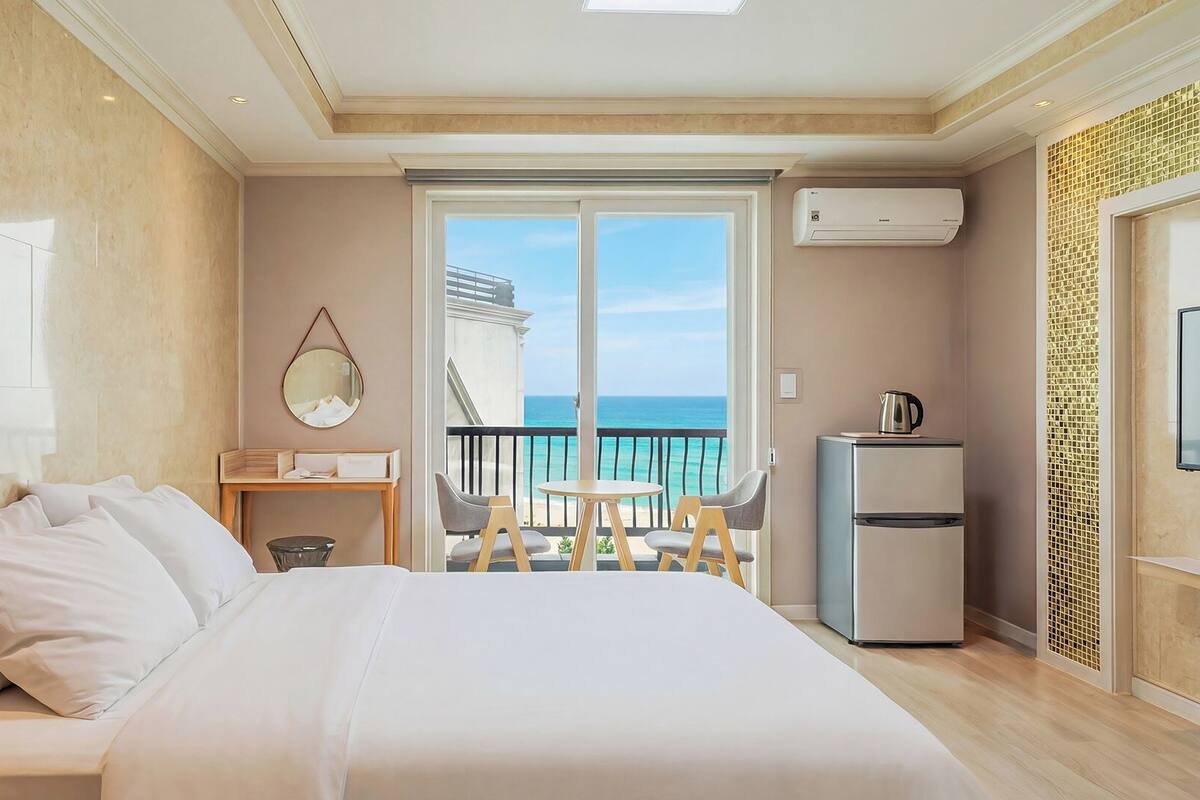 Une chambre d’hôtel comprenant un lit, une petite table, une chaise, un réfrigérateur et un balcon avec vue sur la mer.