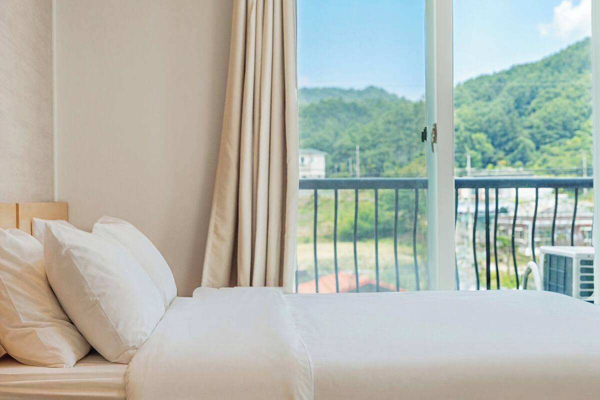 Une chambre d’hôtel avec un lit, des oreillers blancs et une vue sur la verdure et les bâtiments, visible depuis un balcon.