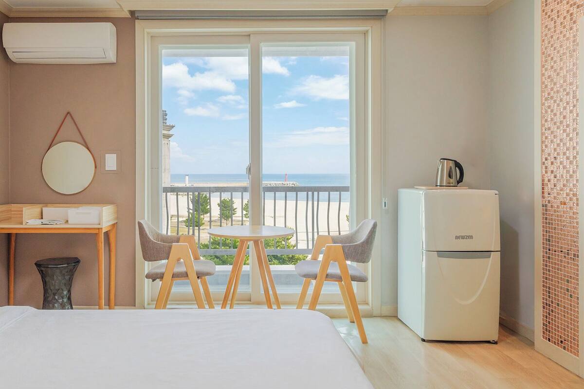 Une chambre d’hôtel avec un lit, une petite table avec des chaises, un réfrigérateur, un miroir et une vue sur la plage.