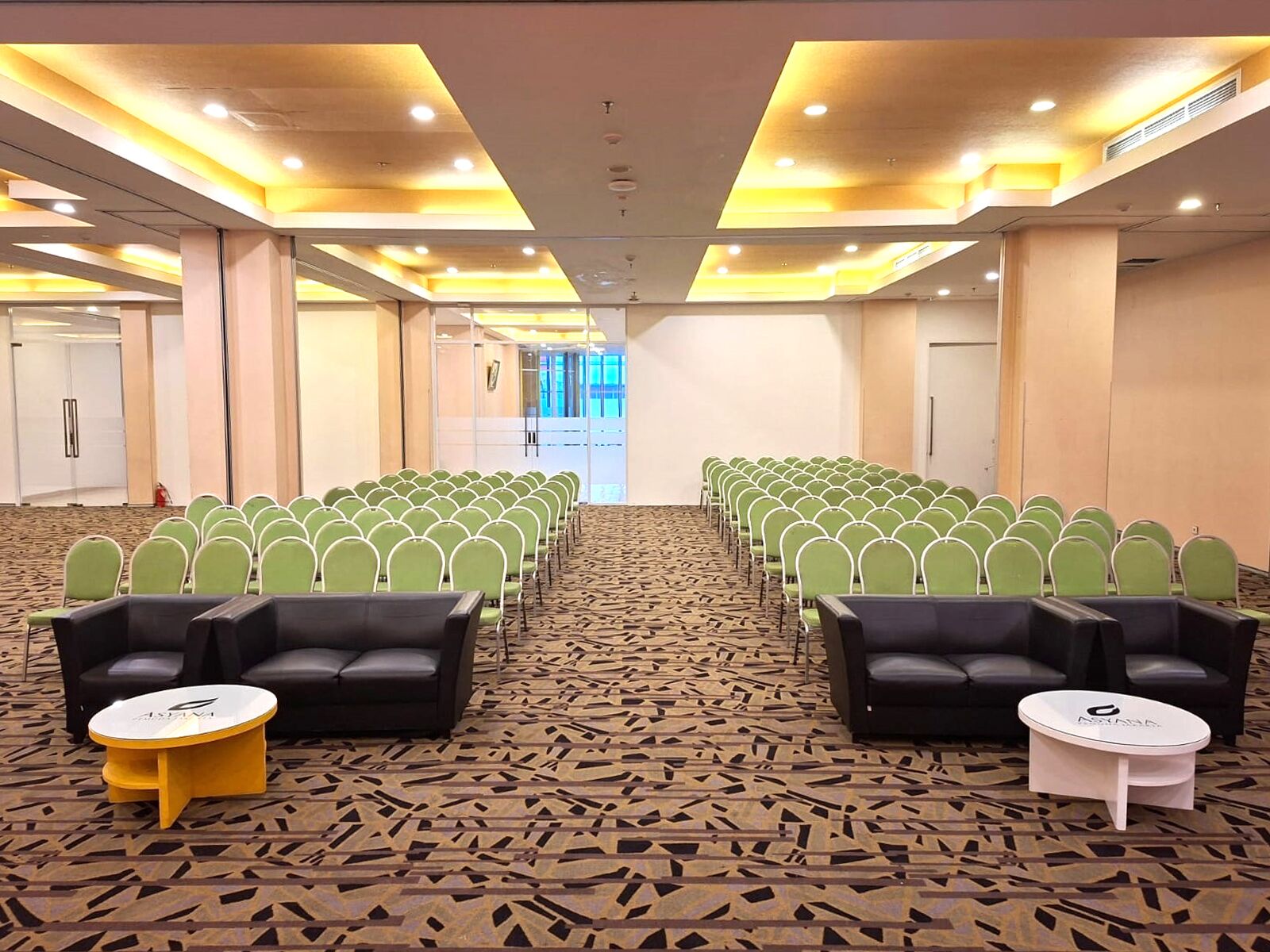 Ein Konferenzraum mit Reihen grüner Stühle, schwarzen Sofas und kleinen Tischen, mit Teppichboden und hoher Decke mit eingelassener Beleuchtung.