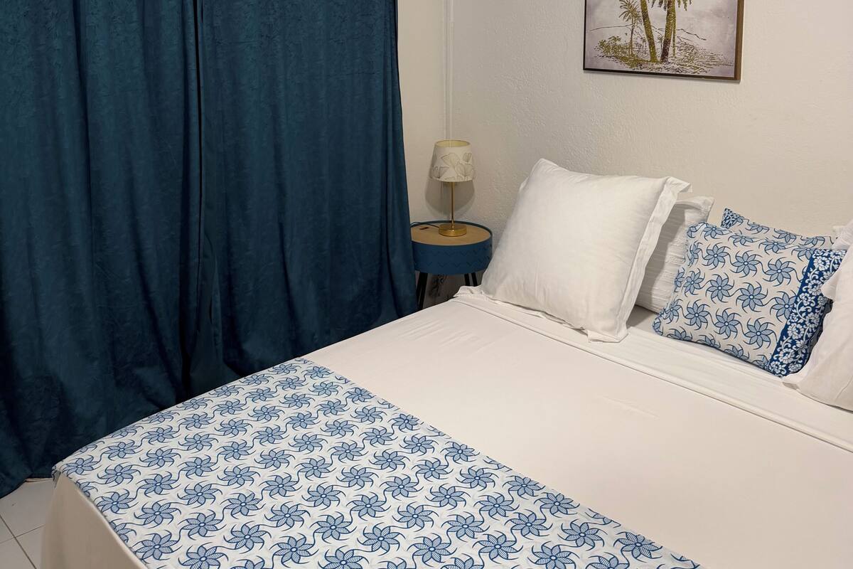 A bedroom with a bed, curtains, a wall-mounted air conditioner, a bedside lamp, and a framed picture.