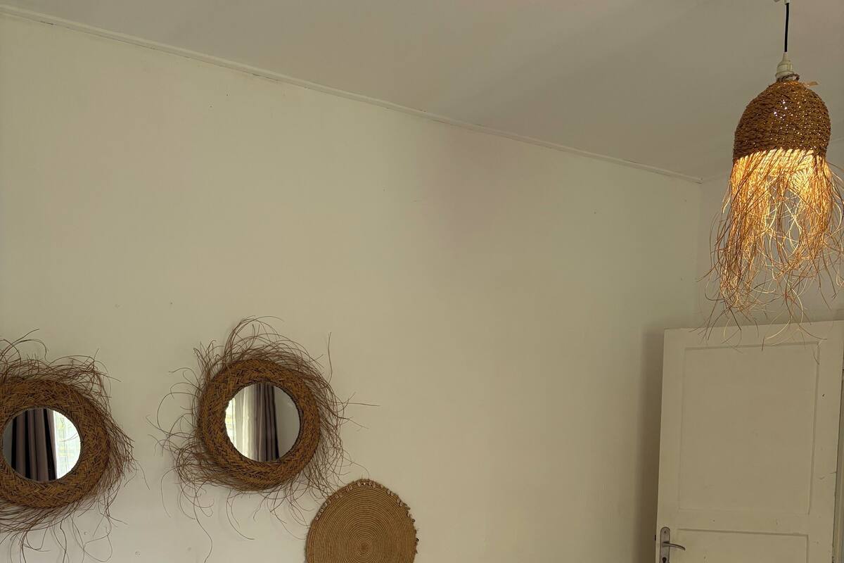 A bedroom with a bed, two wall-mounted mirrors, a woven wall decoration, a hanging light fixture, and a white wardrobe.