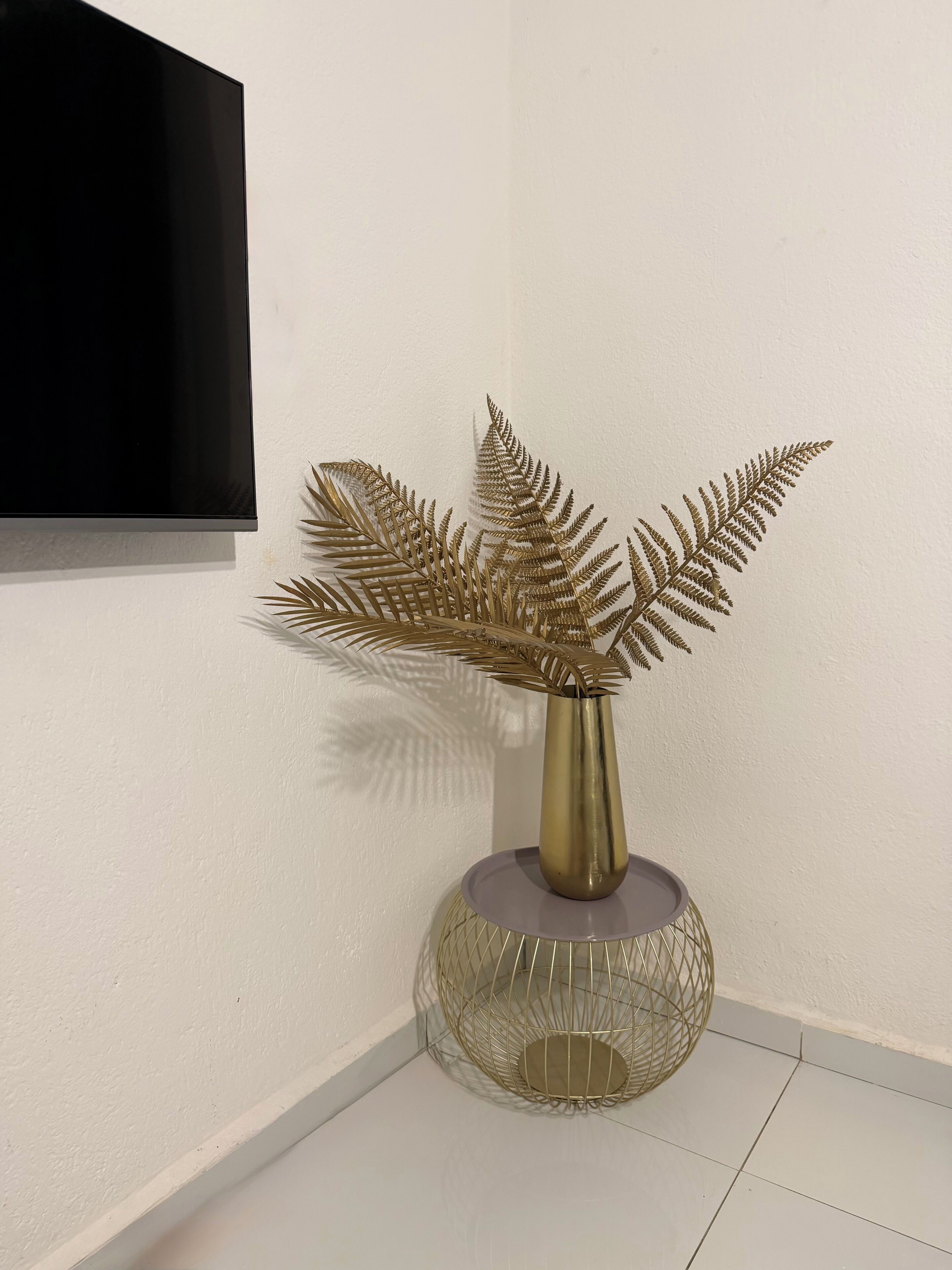 Une plante décorative de couleur dorée, placée dans un vase sur un socle en fil de fer, est posée sur un sol carrelé blanc, devant un mur uni.