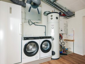 Ein Waschraum mit zwei Waschmaschinen, einem großen Warmwasserbereiter sowie sichtbarer Sanitär- und Elektroinstallation.
