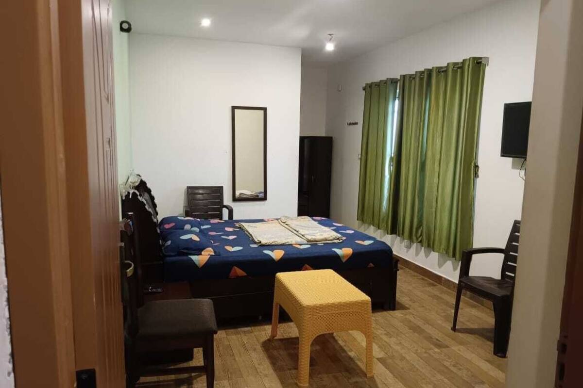 Une chambre à coucher comprenant un lit, une chaise, une petite table, un miroir, un téléviseur et une fenêtre avec des rideaux.