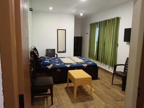 Une chambre à coucher comprenant un lit, une chaise, une petite table, un miroir, un téléviseur et une fenêtre avec des rideaux.