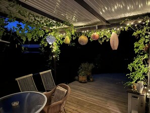 Une terrasse extérieure aménagée avec des meubles en osier, des guirlandes lumineuses et des lanternes suspendues.