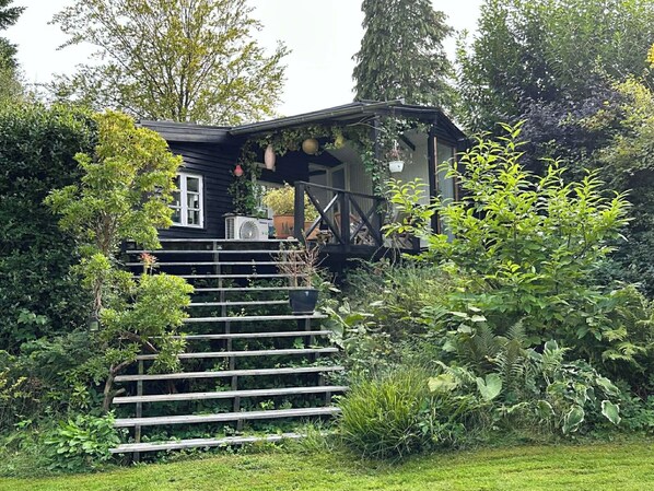 Une maison en bois dotée d’un porche, entourée d’une végétation luxuriante, et un escalier en bois menant à l’entrée.