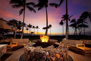 A stone fire pit with a fire in the center, surrounded by wicker furniture, set against a sunset backdrop with palm trees. - Beachfront Westin Kā‘anapali Resort + Amenities . STUDIO (Lahaina)