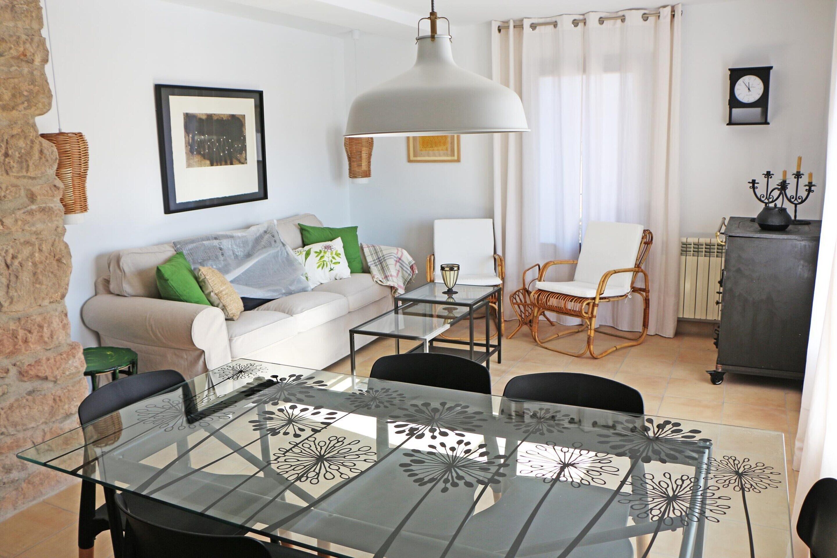 A living room with a sofa, chairs, a glass dining table, a hanging lamp, and a clock on the wall.