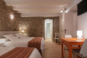 Habitació amb parets de pedra, un llit amb llençols blancs, una taula de nit amb una làmpada i una taula de fusta amb una cadira.