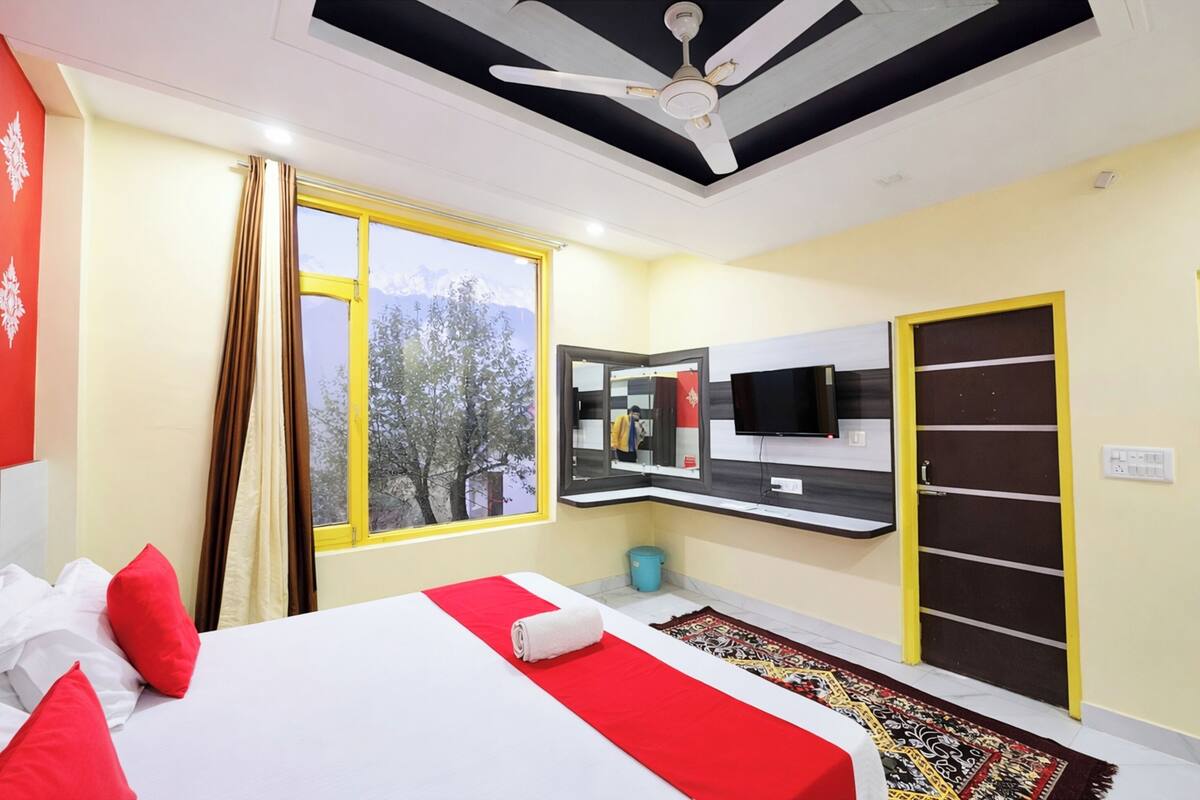 Een hotelkamer met een bed, een plafondventilator, een raam met gordijnen, een spiegel, een televisie en een deur.