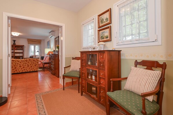 Una habitación con un armario de madera, un sillón tapizado en verde, una alfombra estampada y un sofá al fondo.