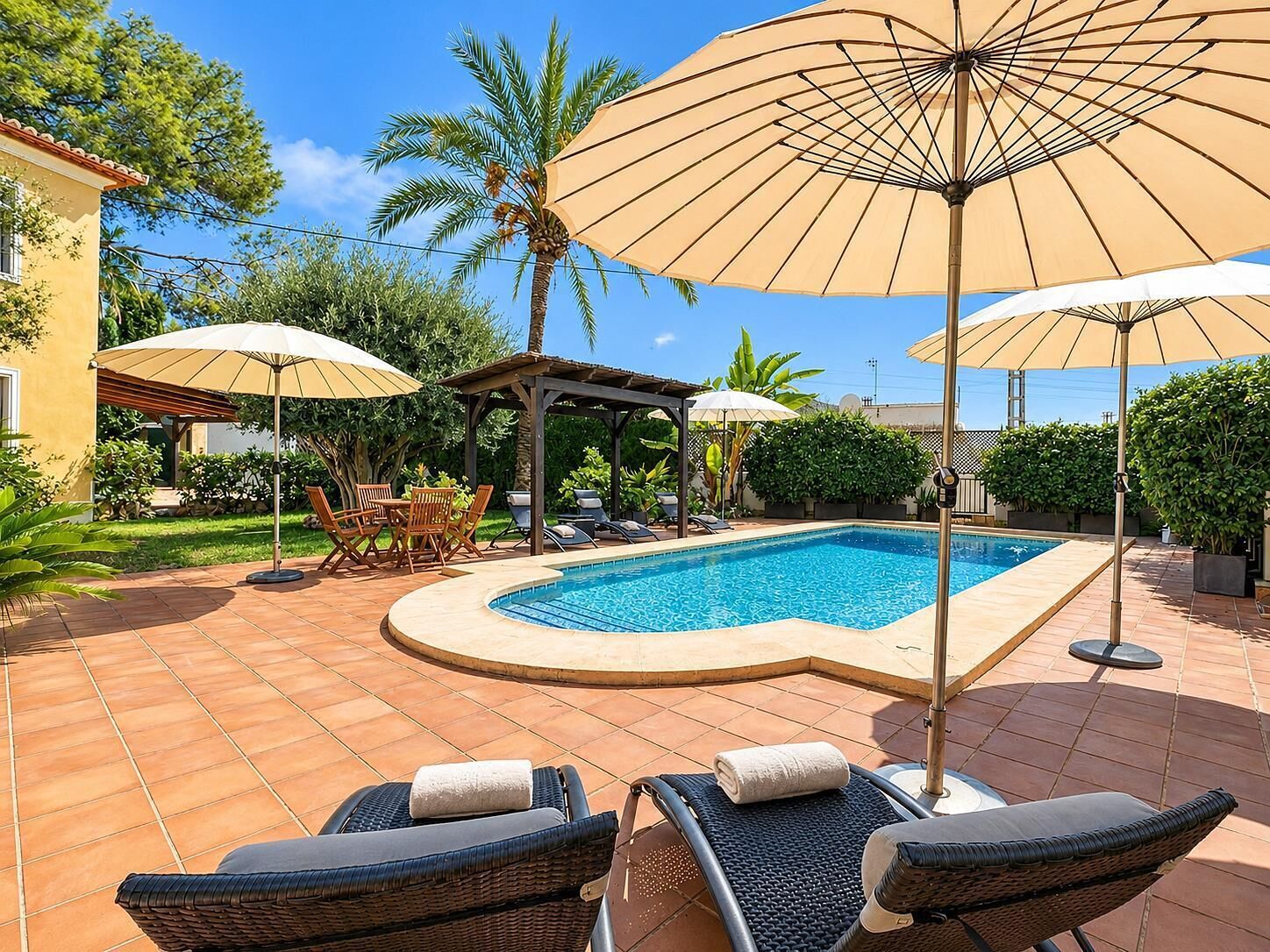 A swimming pool with lounge chairs and umbrellas, surrounded by a garden and a building.
