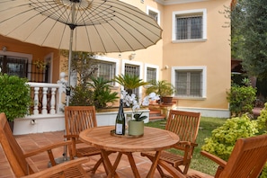 Une terrasse extérieure avec une table ronde en bois, quatre chaises en bois, un parasol beige et des plantes en pot.