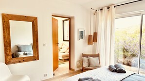 Une chambre avec un grand lit, un miroir à cadre en bois, une chaise blanche et une vue sur un jardin, accessible par une porte coulissante.