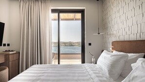 A hotel room with a bed, a TV, a wooden nightstand, a bottle, and a view of the sea. - Infinity Rise Villa, a Distinctive Private Stay (Agia Pelagia)
