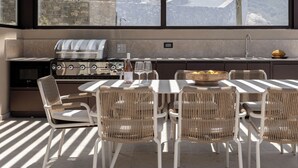 An outdoor kitchen with a grill, dining table, and chairs, overlooking a mountainous landscape. - Infinity Rise Villa, a Distinctive Private Stay (Agia Pelagia)