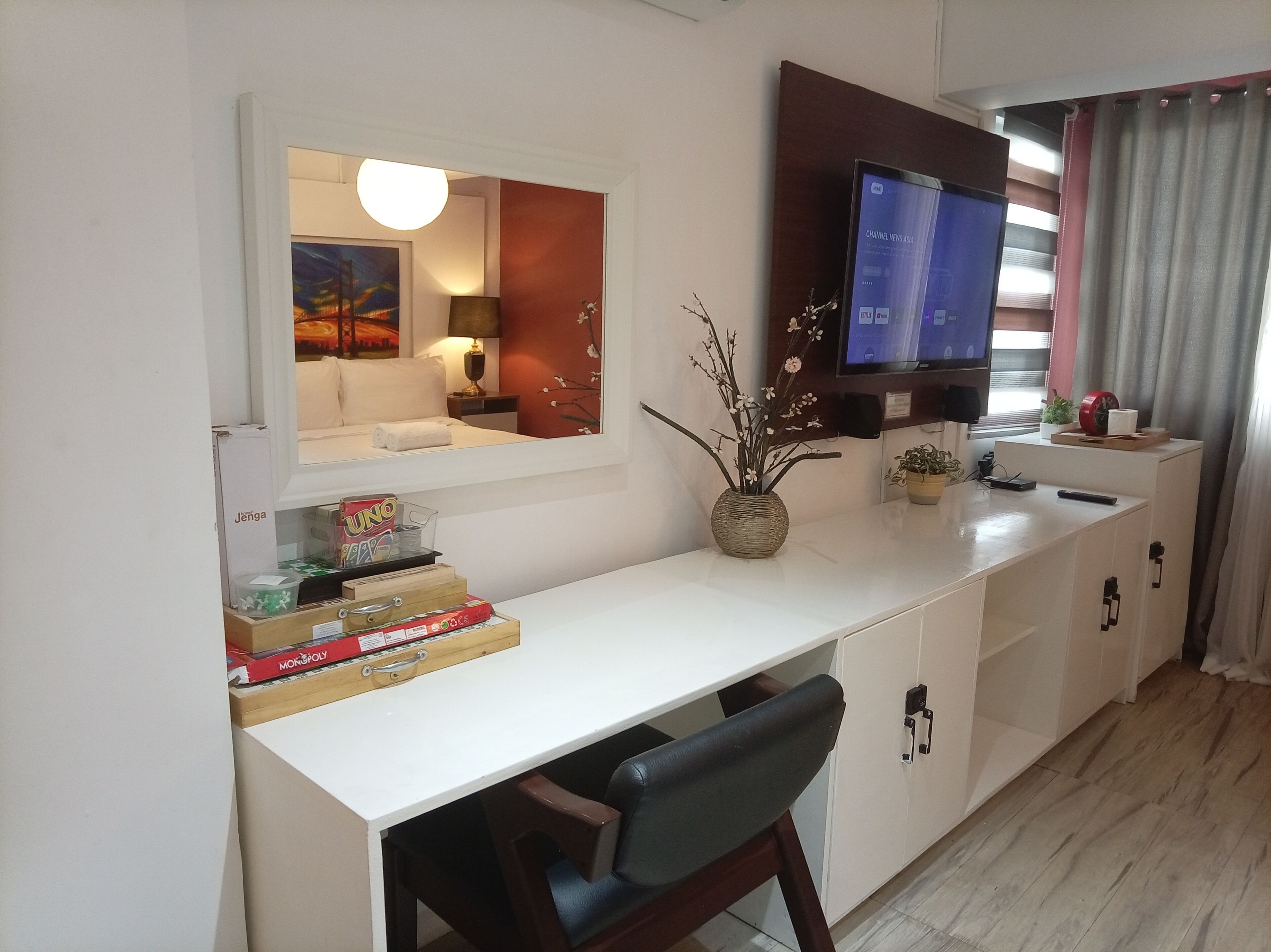 A hotel room with a white desk, a chair, a TV, a bed, a mirror, and a window with curtains.
