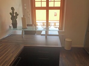 A room with a glass table, a potted cactus, a small plant, a white trash bin, and a window with a view of buildings. - Apartment 100 sqm separate bedroom and balcony, App.9 (Aschau im Chiemgau)