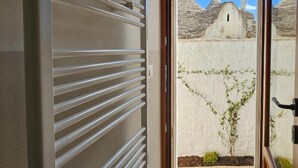 Vista a través de una puerta de metal con barras horizontales, que da a un patio con una pared blanca, un árbol pequeño y un edificio de piedra con techo cónico al fondo.