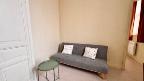 Een kamer met een grijze bank, twee bloemenkussens, een kleine groene tafel en een witte radiator.