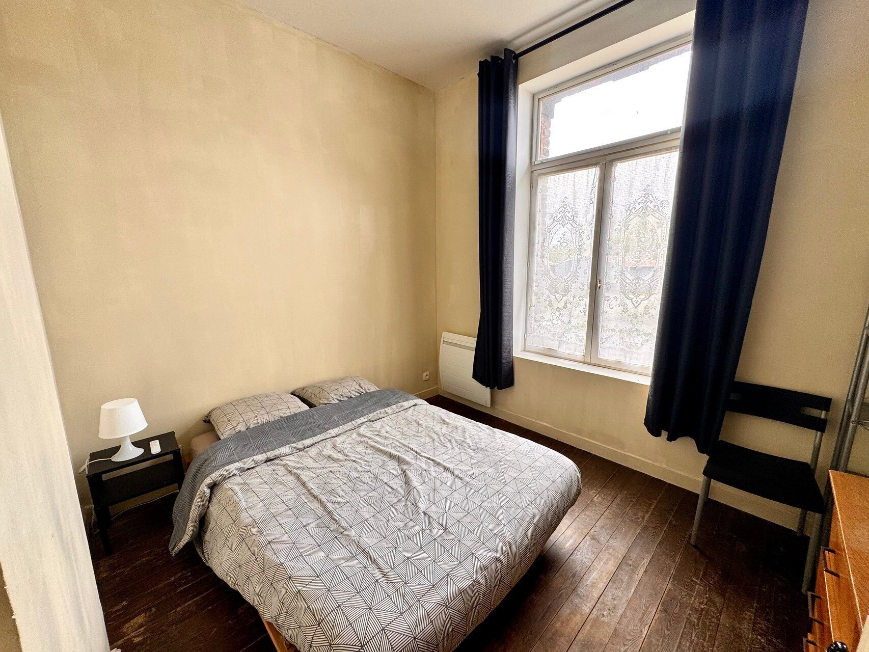 A bedroom with a bed, a nightstand, a chair, a window with curtains, and a radiator.