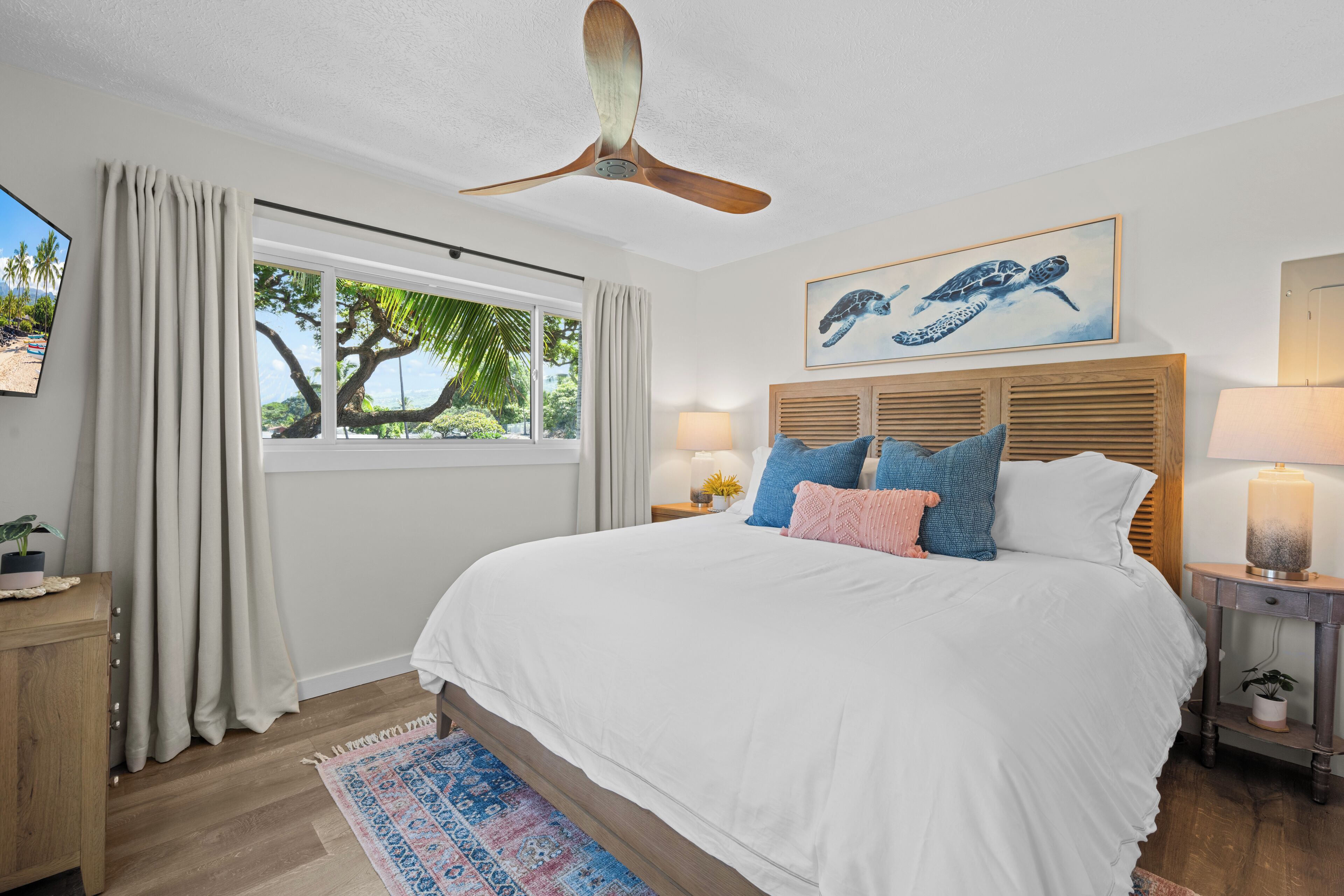 A bedroom with a bed, a ceiling fan, a window with curtains, a nightstand with a lamp, and a painting of a sea turtle.