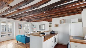 A modern kitchen with exposed wooden beams, a blue sofa, and a large window. - Appartement 2 pièces à proximité du marais (Paris)