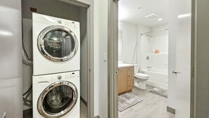 Une buanderie équipée de deux machines à laver à chargement frontal, d’un meuble en bois et d’un espace douche comprenant une baignoire et des toilettes.