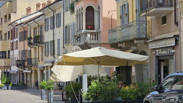 A street with multi-story buildings, outdoor seating with umbrellas, and a car parked on the side. - Elegant apartment in the heart of Vigevano, a stone's throw from Piazza Ducale (Vigevano)