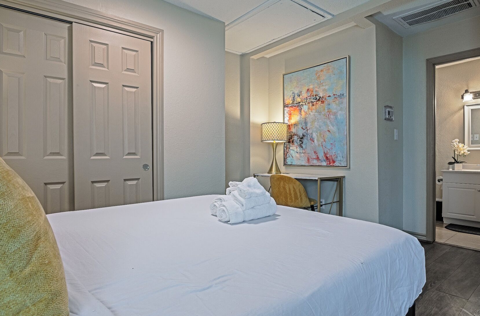 A hotel room with a bed, a desk, a chair, a painting, a lamp, and a bathroom.