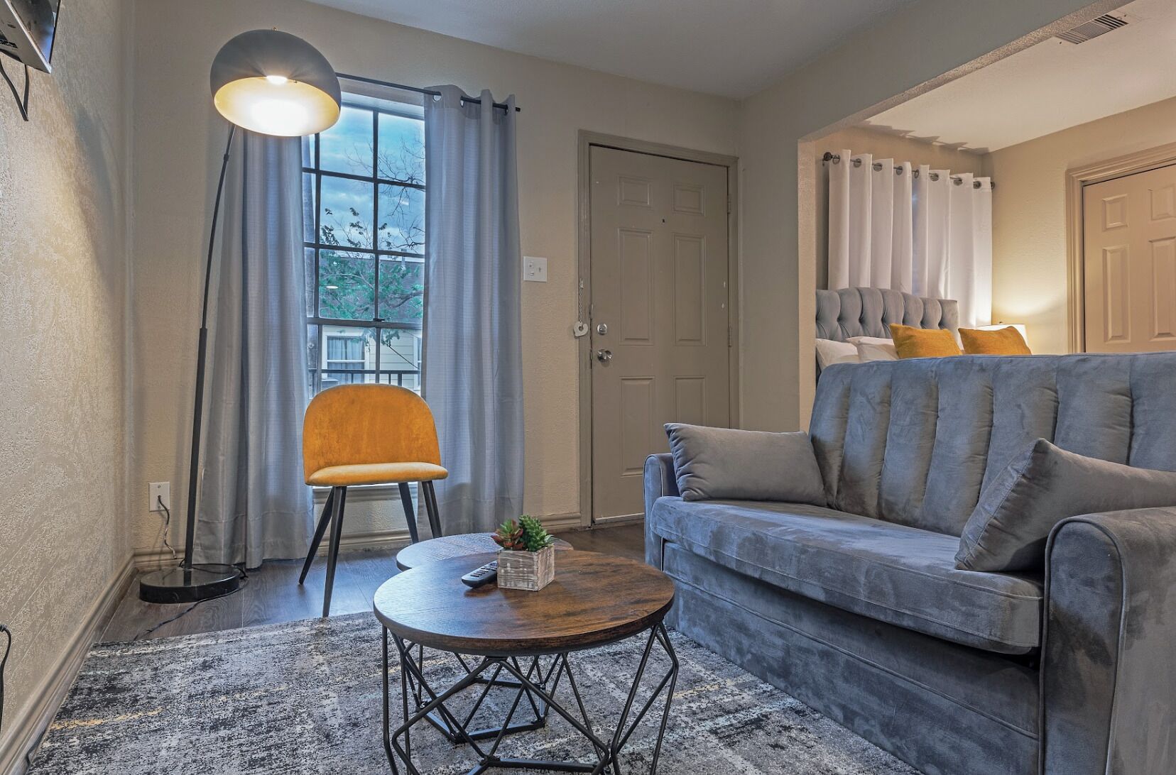 Un salon comprenant un canapé, un fauteuil, une table basse ronde, un lampadaire et une fenêtre avec des rideaux.