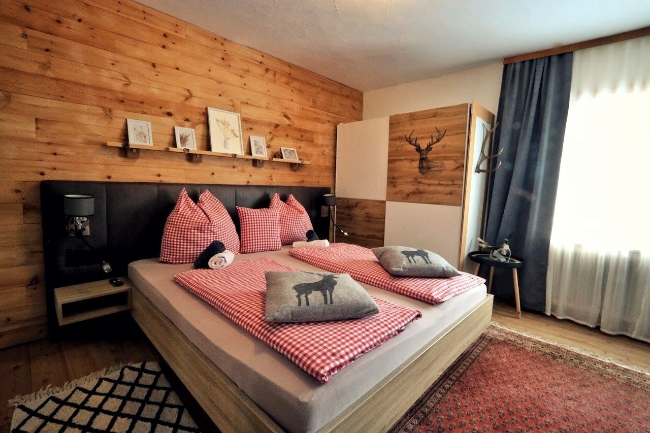 Ein Schlafzimmer mit Holzwand, einem Bett mit karierter Bettwäsche, einem Kopfteil, Nachttischen und einem aufgehängten Hirschkopf.