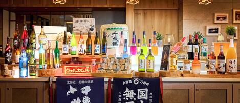 Zona de bar con una variedad de botellas, incluyendo licores y sake. Hay armarios de madera y un cartel con caracteres japoneses.