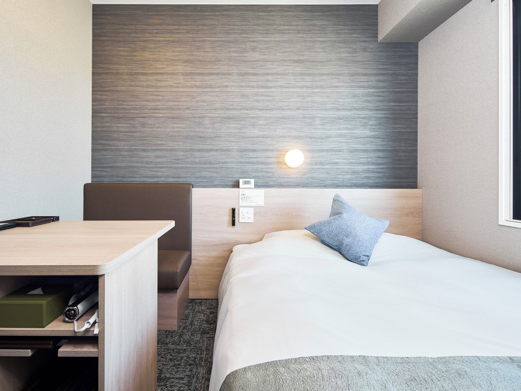 Une chambre d’hôtel avec un lit, un bureau, une chaise, une lampe fixée au mur et une fenêtre.