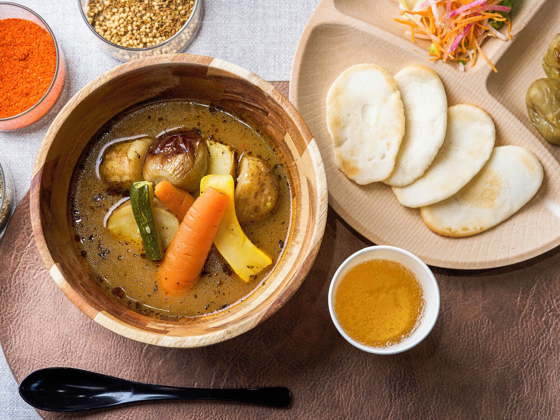 A wooden bowl with a stew containing vegetables and meat, a plate with flatbreads, and a small bowl of sauce.