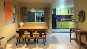 Un comedor con mesa y sillas de madera, una ventana con cortinas, un ventilador y vistas a una cocina con armarios verdes.