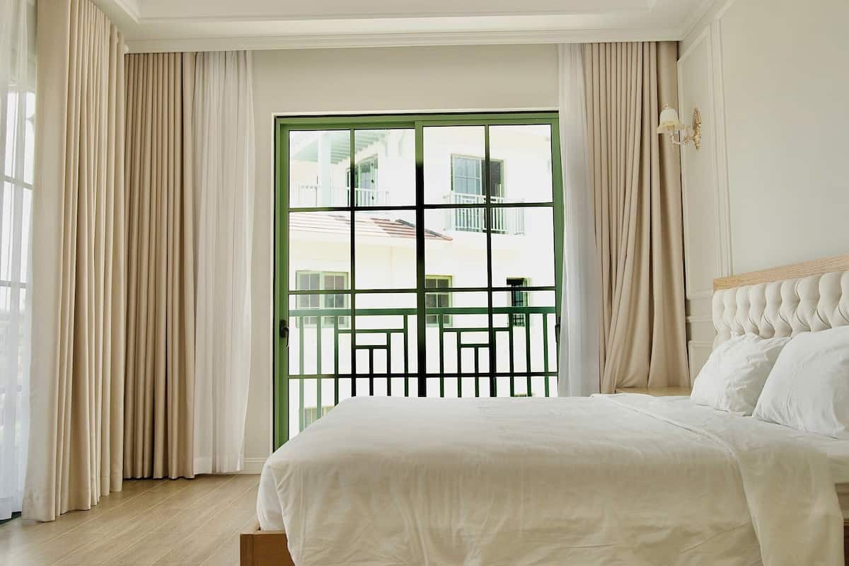 A hotel room with a bed, curtains, a window with a view, and a wall-mounted light fixture.
